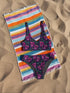 Two-piece bikini set featuring a bold pastel goth design with neon pink skulls wearing green bows, surrounded by multicolored stars on a deep black background. The set includes a plunging V-neck bikini top with wide straps and high-waisted bottoms for a flattering retro fit. Made of stretchy, soft material with a smooth black lining. This skull print swimsuit is ideal for alternative, edgy summer style with a playful punk twist.
